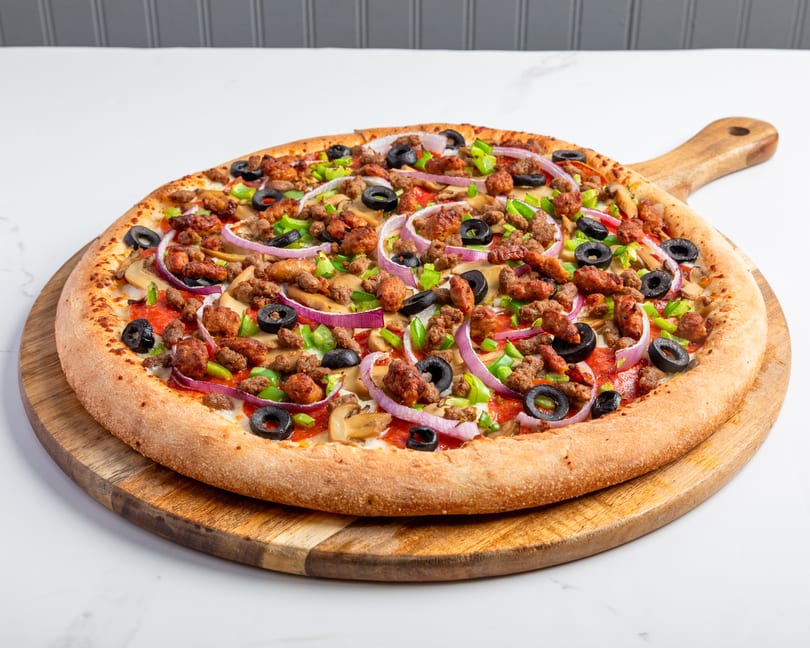 A fully loaded pizza with sausage, pepperoni, olives, onions, and peppers on a wooden serving board.