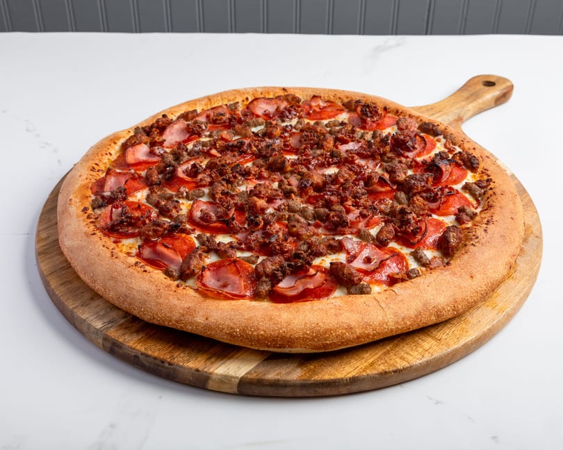 A large, round meat lover's pizza with pepperoni, sausage, and ham on a wooden paddle.