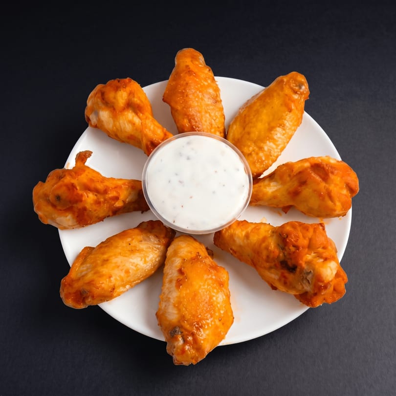 Seven buffalo chicken wings arranged around a small cup of ranch dressing on a white plate.