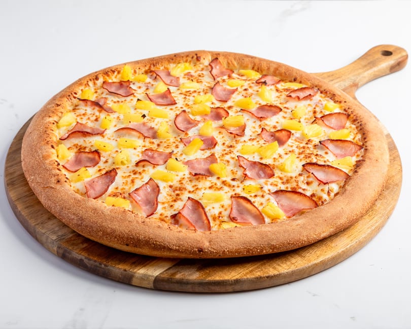 Hawaiian pizza with ham and pineapple on a wooden serving board.