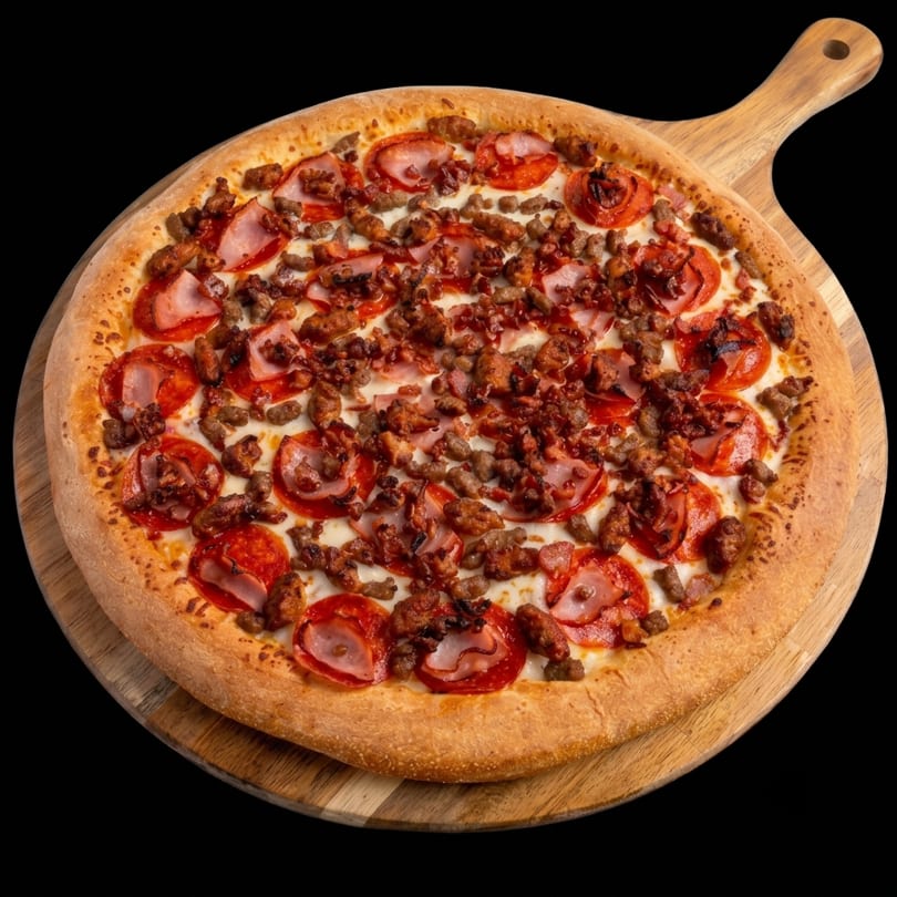 A large, meat-lover's pizza with pepperoni, ham, and sausage on a wooden pizza peel.