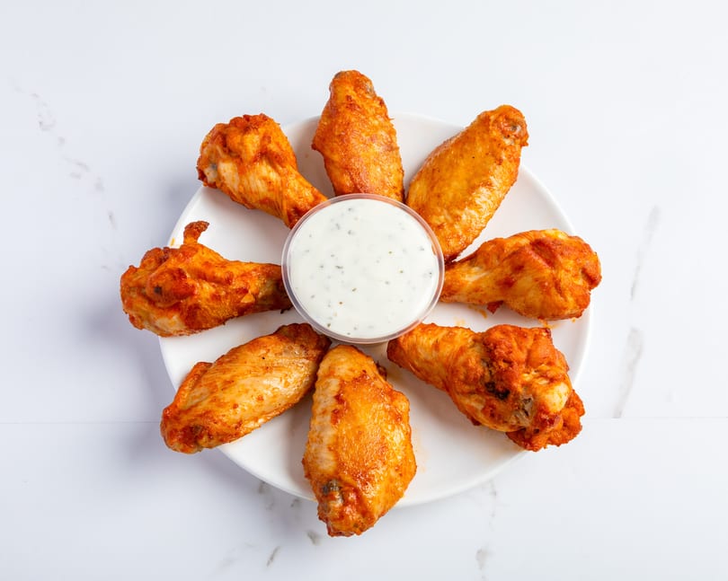 Seven spicy chicken wings arranged in a circle around a small cup of ranch dressing on a white plate.
