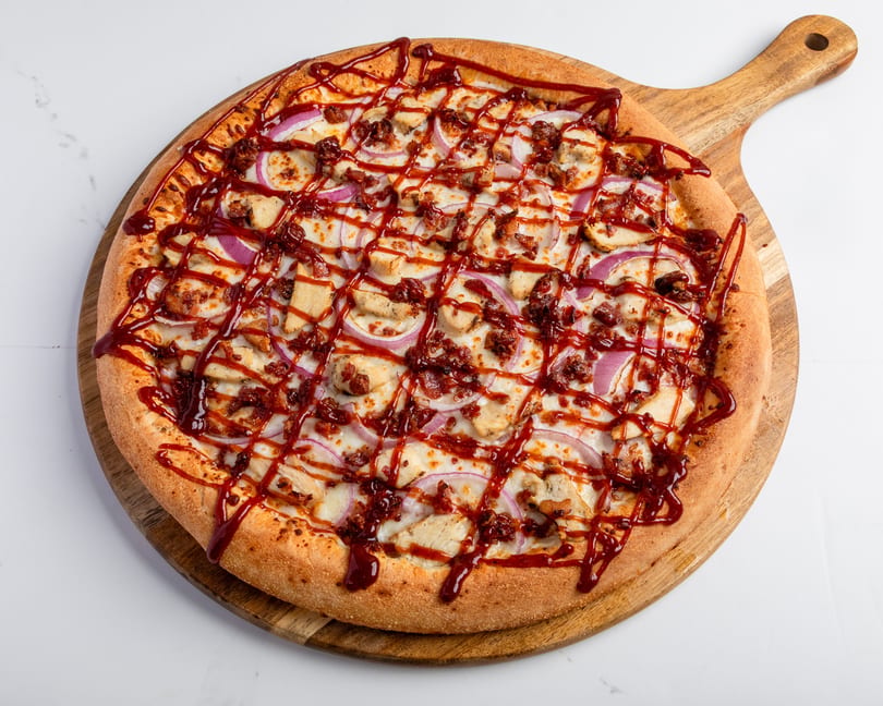 BBQ chicken pizza with red onions and bacon, drizzled with BBQ sauce on a wooden peel.