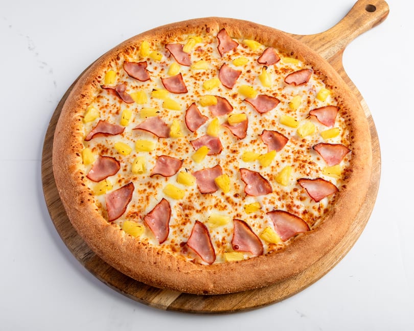 Hawaiian pizza with ham and pineapple on a wooden pizza peel, against a white marble background.